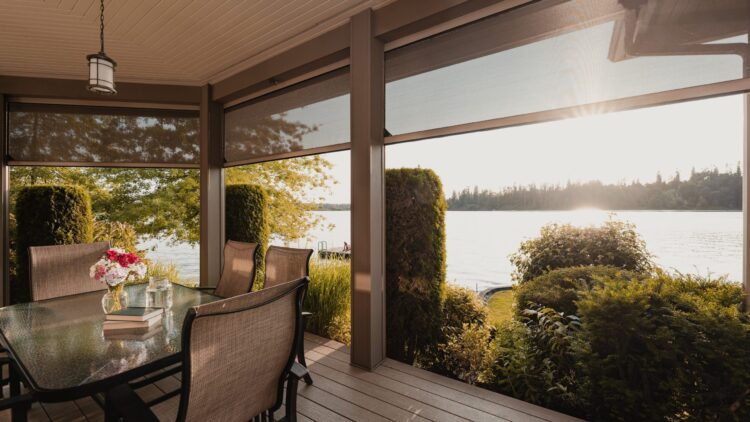 Motorized patio screens on a covered porch overlooking a lake at sunset, with outdoor dining furniture and lush landscaping