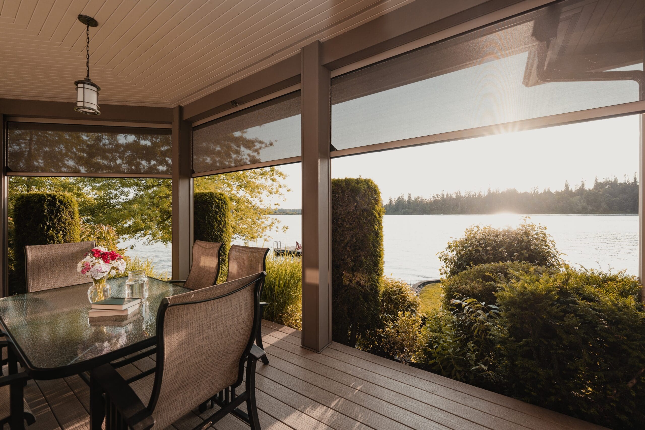 Motorized patio screens on a covered porch overlooking a lake at sunset, with outdoor dining furniture and lush landscaping