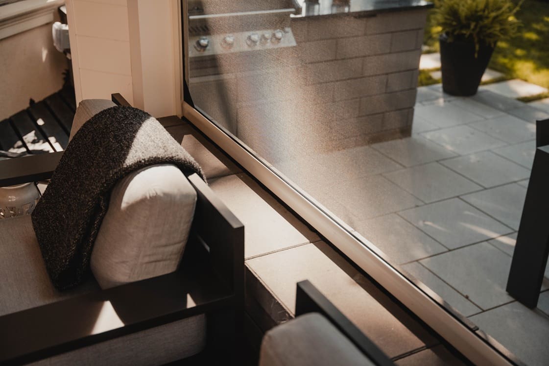 Close-up of a patio seating area with a cushioned chair, dark throw blanket, and motorized screen casting shade on a tiled outdoor floor